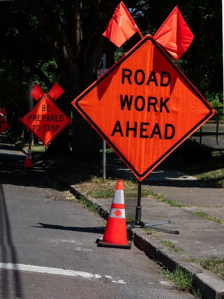 Orange road work ahead sign and cone on city street in Vancouver, WA, under sunny conditions.